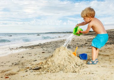 Happy little blond boy play with sand pouring water from plastic bucket on the sea beach during vacation view from profile
