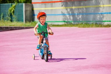 Little blond boy ride small bicycle with learning wheels on color surface of the park