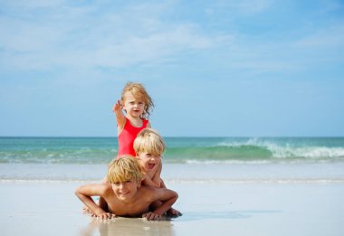 Group of three happy children lay on top of another on the sea sand beach at summer vacation with girl point finger