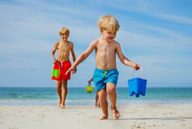 Little blond boy with other kids on background run carrying toy bucket playing on sea sand beach