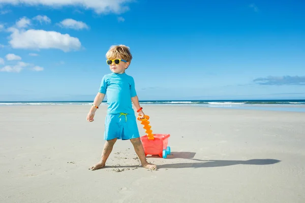 Happy little boy in sunglasses walk on the white sand beach playing with toy cart