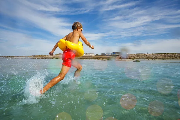 Young boy in orange sunglasses run on the beach at the sea with inflatable duck view from behind