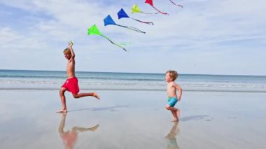 Two handsome brother boys run together holding colorful  kite over blue sky at the ocean beach having fun on summer vacation