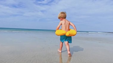 Happy little blond boy with inflatable yellow duck buoy run on the sand beach