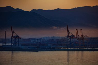 Akdeniz 'de Akdeniz körfezinde, Malaga şehri ve dağları üzerinde vinçleri olan endüstriyel bir kargo terminalinin gün batımı görüntüsü