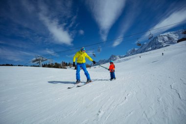 Adult on skiing slope glide downhill teaching little child to ski both holding poles clipart