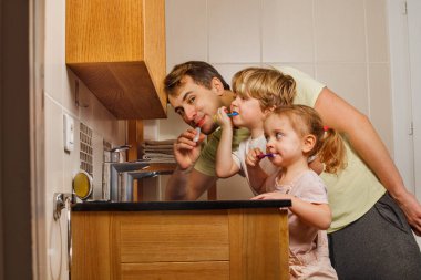 Aile babası, iki küçük çocuk, banyoda diş fırçası kullanan kız diş fırçası, ağızda fırça ve kameraya bak.