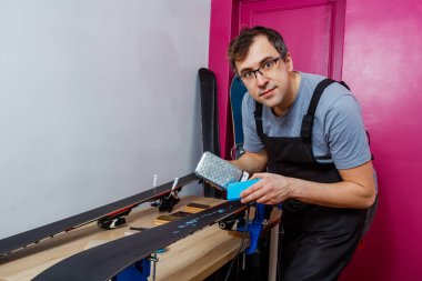 Atölye çalışanı kayak tabanına demir ağda kayakları kullanarak mum damlatıyor ve kameraya bakıyor.