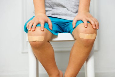 Küçük bir çocuğun dizine yakın çekim, üzerinde bir çiziği kapatan bir yama var, eller dizlerinin üzerinde yatıyor.