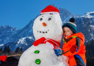 Kışın spor kıyafetli şirin bir çocuğun Noel Baba şapkalı bir kardan adamla oynadığı yakın plan portresi.