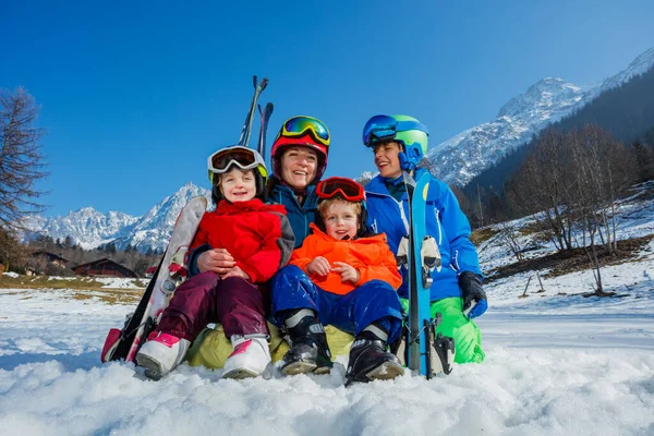 Family, mother son and little kids sit in the snow in sport outfit with skies helmets masks laugh hug smiling over mountain
