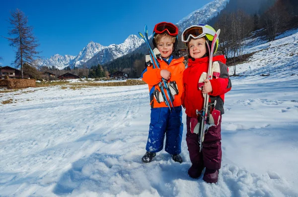 Two kids children and girl at their first alpine getaway, wear helmets, ski masks while standing with skis on the snow-covered mountain terrain