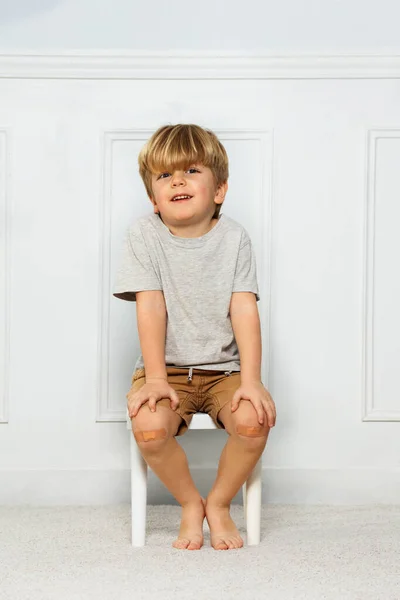 Young boy with knee having patch covering a scratch, while his hands and fingers lay on knees smiling sitting on a stool