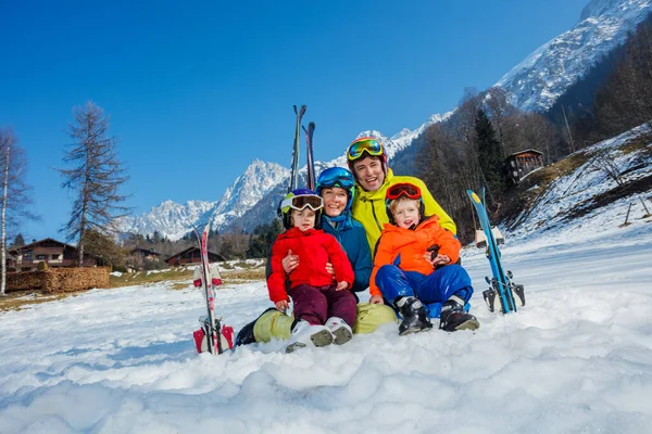 儿子和小孩穿着运动服坐在雪地里 戴着滑雪板 戴着钢盔 笑着拥抱高山 — 图库照片