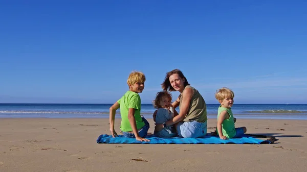 妈妈带着三个金发碧眼的孩子 男孩和女孩坐在海边的垫子上 在阳光明媚的日子回头看 — 图库照片