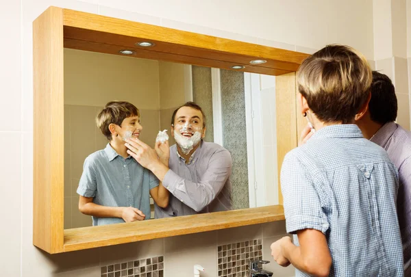 Father with kid during funny bonding shaving and foaming, each other in bathroom, smiling, view through the mirror