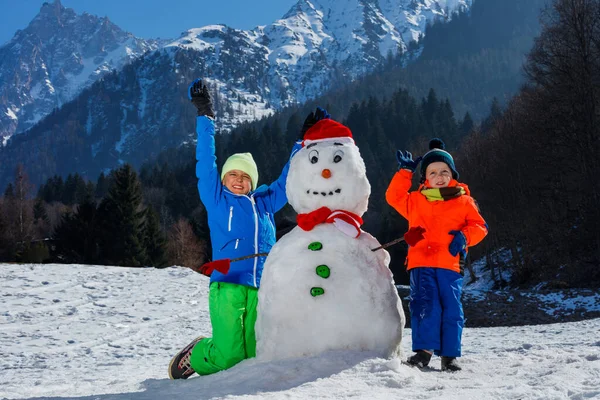 两个穿着冬季运动服的快乐男孩兄弟在山外与戴着圣诞礼帽的雪人玩耍 — 图库照片