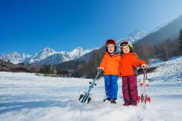 Two kids boy and girl at their first alpine getaway, wear helmets, ski masks while standing with skis on the snow-covered mountain terrain