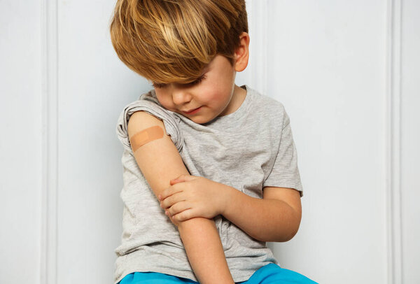 Portrait of a calm handsome little blond boy with band-aid on his shoulder touching hand looking at scratch
