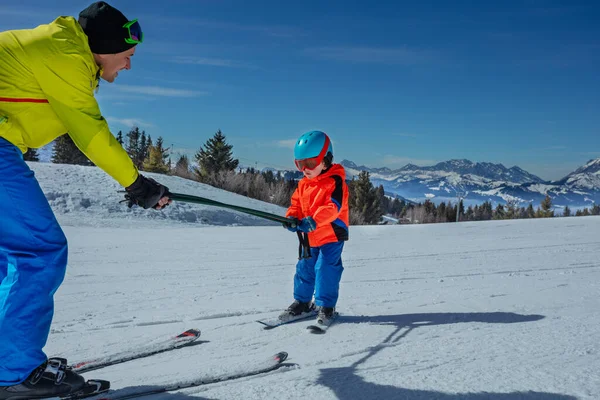 男は下に転がって棒を握ることによってアルプスの斜面でスキーするために小さな少年を教えます — ストック写真