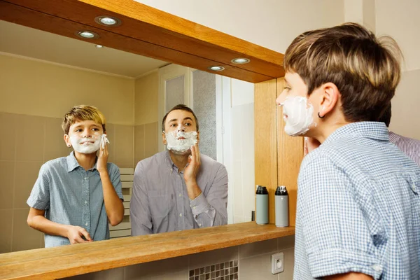 Father and the son have fun in the bathroom shaving putting foam on the face looking at the mirror