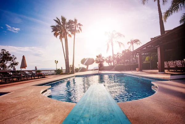 从跳水板后面看到游泳池 日光浴床和遮阳伞 阳光透过棕榈树的叶子照射着温暖的落日 — 图库照片