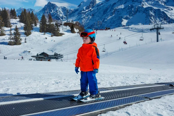 マスクとヘルメットが付いている小さな男の子は 登山トップ上の冬の眺めの間にスキースクールで歩道ベルトを移動します — ストック写真