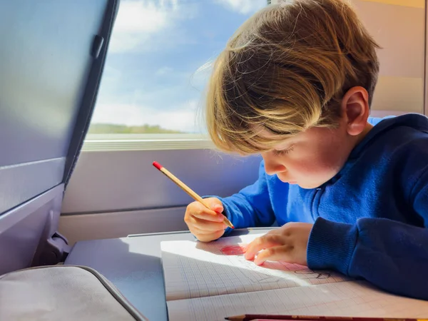 小さな男の子はノートパッドの鉛筆で書く高速列車の中の窓に座っています — ストック写真