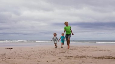 Yaz kıyafetli çocuklar kumsalda el ele tutuşup gülümsüyorlar.