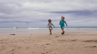 Oğlan ve kız, yaz kıyafetleri içinde iki küçük çocuk kumsalda birlikte koşuyor, gülümsüyor ve eğleniyorlar.