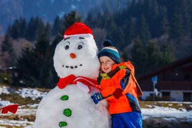 Kışın spor kıyafetli mutlu küçük bir çocuğun yakın plan portresi dışarıda Noel Baba şapkalı bir kardanadamla oynuyor.