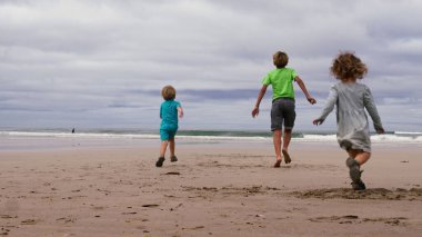 Üç çocuğun arkasından, yaz kıyafeti giymiş küçük bir kız ve erkek çocuğu kumsalda birlikte koşuyor ve eğleniyorlar.