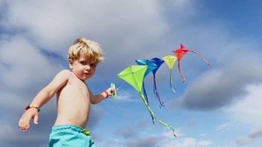 Close-up of a little boy smile, laugh stand holding many kites in hand and looking at them smiling over blue sky