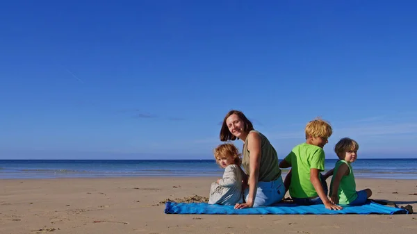 Family of mom with three blond kids boys and girl sit on the beach mat at the ocean turning back at sunny day