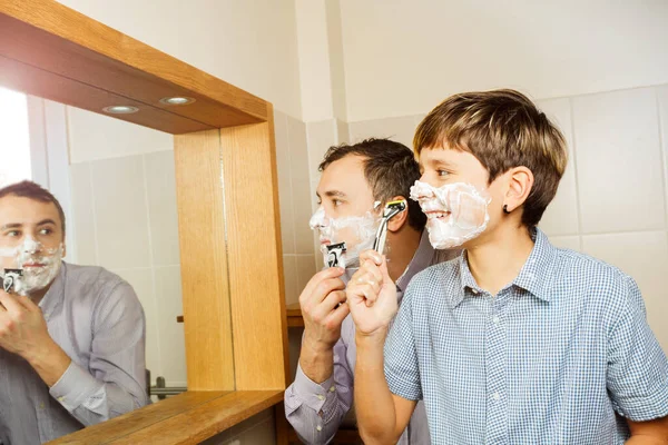 Father and the son have fun in the bathroom shaving together looking in the mirror smiling wearing shirt
