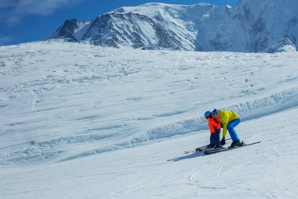 父親は雪だるまの動きのプロフィールビューを示す手でスキーバインディングの後ろにスキーグライディングに小さな子供を教えます — ストック写真
