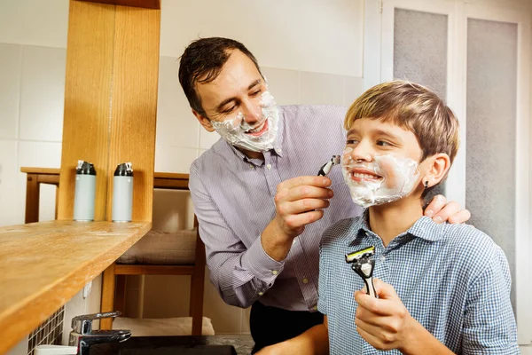Dad shows and teaches son how to shave, holding razor with foam on boy\'s cheek, smiling having fun