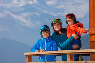 Balkonda kayak kasklı bir aile, Alp Panorama 'da gülümsüyor, anne küçük oğlunu kollarına alıyor.