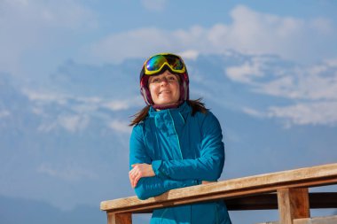 Ahşap balkonda dikilen mutlu kadın kameraya bakıyor miğfer, maske ve kayak ceketi giyiyor.