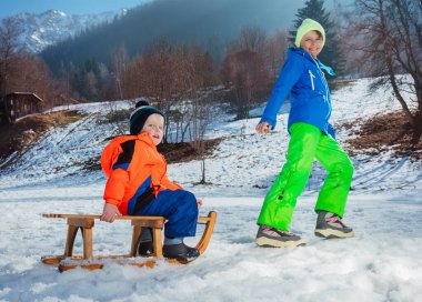 Kardeş yokuş yukarı, yüksek dağların tepesindeki tahta kızakta bir çocuk ve kışın kardan adam çekiyor.