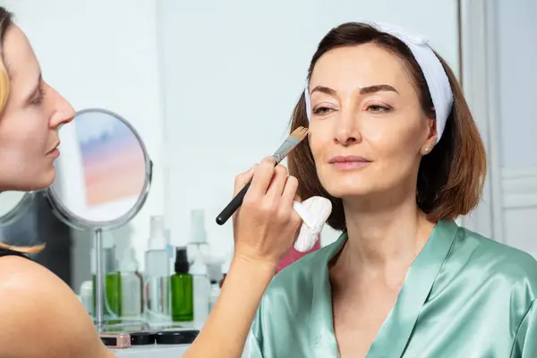 美容师在中年妇女的脸上涂上主要的粉底霜 并在镜子前戴上头巾 — 图库照片
