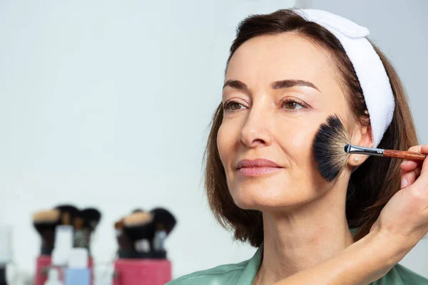 Beautician using fan makeup brush on the face of middle aged woman wearing headband sitting before mirror