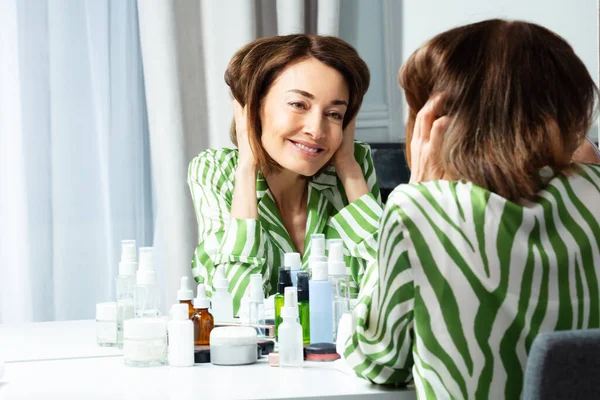 一名中年妇女穿着绿色长袖长袍 在浴室梳妆镜里摸头发的画像 — 图库照片