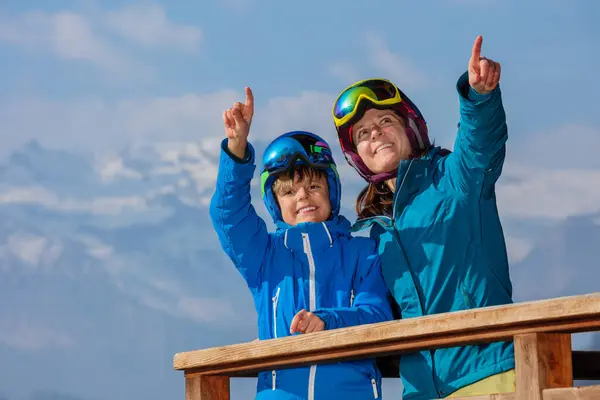 Smiling ski family on chalet balcony admiring Alps in helmet and mask overlooking mountains, talking showing slopes