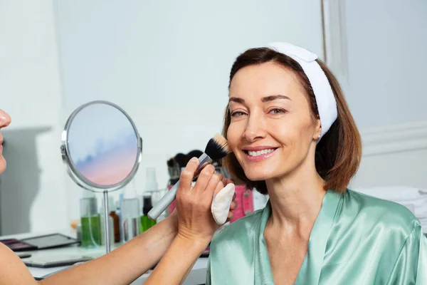 大きなブラシを使用した化粧を施した美容師との幸せな広い笑顔の中年女性の肖像 — ストック写真