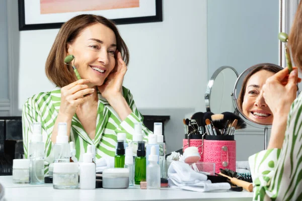 浴室里穿着长袖衣服的中年妇女的微笑肖像在镜子前使用面部滚筒进行皮肤护理 — 图库照片