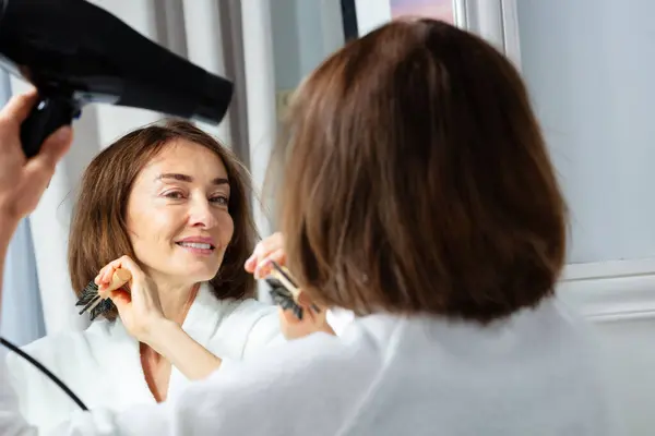 化粧鏡の前にヘアドライヤードライヤーの髪を保持するハウスコートの美しい中年の女性のクローズアップ美しさの肖像画 — ストック写真