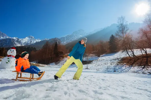 一个母亲在高山上拖着一个男孩爬上木制雪橇 冬天又拖着雪人的照片 — 图库照片