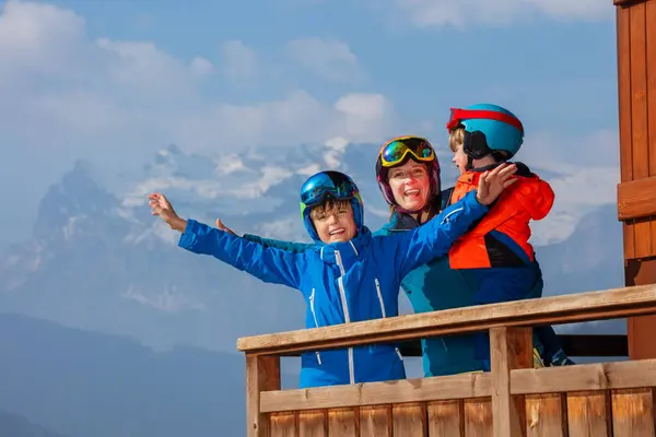 Family in ski helmets on balcony, smiling over Alpine panorama, mother hold little skier in her arm, happy older son raising arms in freedom gesture