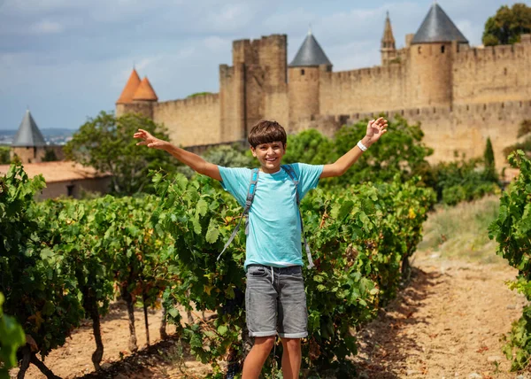 ブドウ畑に立っているバックパックとTシャツでハッピーティーン旅行者は手を挙げ フランス フランス 夏休みのコンセプトで壮大なカルカソンヌフランスの要塞都市を見ます — ストック写真